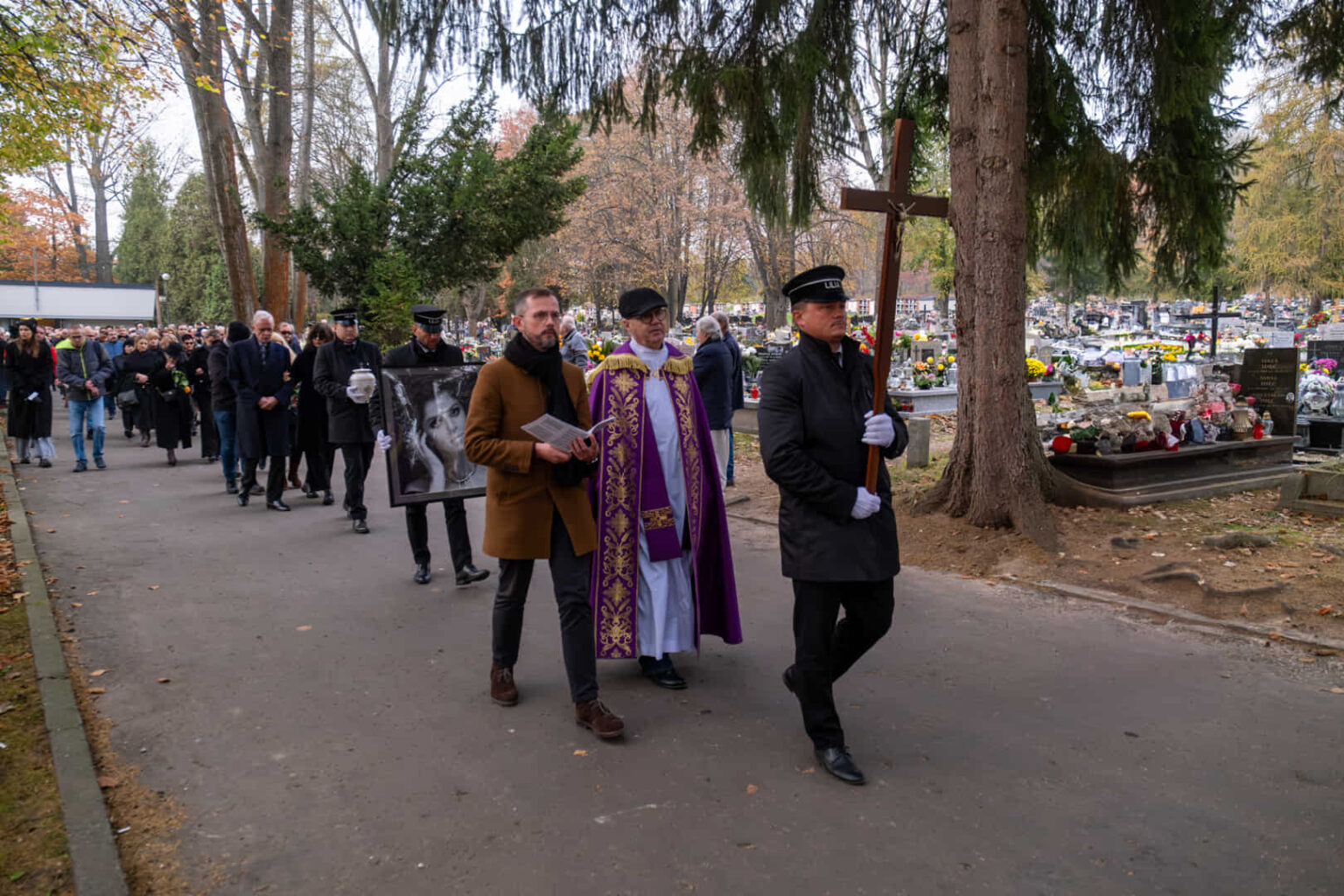 A funeral procession with a priest and a cross in a cemetery avenue